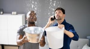 Two men indoors catching heavy water leaks from the ceiling using buckets, one making an urgent phone call – emergency roof leak situation.